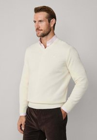 Ecrufarbener Pullover mit V-Ausschnitt aus weichem Material, mit gerippten Bündchen und Saum, getragen über einem hellrosa Hemd mit Kragen und dunklen Hosen.