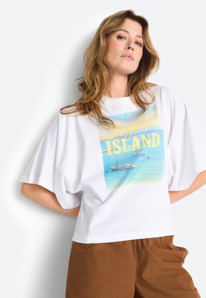 Laineliste juustega naine, kellel on seljas lahtine valge t-särk kirjaga "Lagoon Island" ja purjekate trükiga, kombineeritud pruunide pükstega, positsioneerituna valge tausta ees.