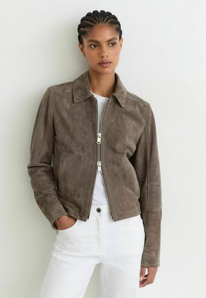Junge Person mit geflochtenem Haar trägt eine braune Wildlederjacke mit Reißverschluss über einem weißen Hemd und weißen Hosen und steht vor einer schlichten weißen Wand.
