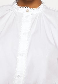 Blouse blanche avec des bords festonnés sur le col et la patte de boutonnage, bouton en perles et strass au niveau du col, portée par une personne.