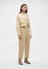 Γυναίκα που φοράει μπεζ cropped σακάκι, αντίστοιχο ψηλόμεσο παντελόνι, ανοιχτόχρωμο πολυεστερικό πουλόβερ με ζιβάγκο και μπεζ μποτάκια στο αστράγαλο, στέκεται μπροστά σε απλό φόντο.