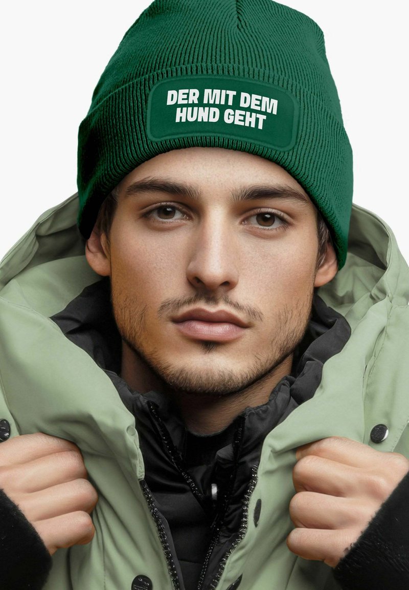 Green knit beanie with white text reading "DER MIT DEM HUND GEHT," worn with a light green jacket and black inner layer, featuring a textured finish.