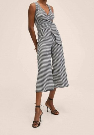 Kobieta w bezrękawowym czarno-białym kombinezonie w kratę, z szerokimi nogawkami i wiązaniem z przodu, w połączeniu z czarnymi sandałami na wysokim obcasie z paskami.