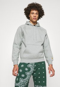 Sudadera gris hecha de tejido suave, con un bolsillo canguro, capucha con cordón y puños acanalados, combinada con pantalones verdes con estampado.