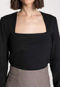 Top nero a maniche lunghe con scollo quadrato, abbinato a una gonna a vita alta con motivo houndstooth nei colori rosso e crema.