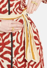 Abito in tessuto con motivo rosso e crema, con vita legata. Presenta una chiusura con bottoni e un accessorio con anello dorato alla mano.