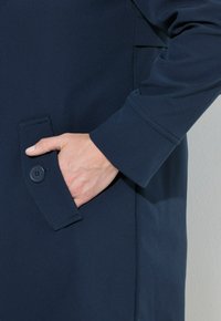 Cappotto blu navy realizzato in un materiale testurizzato, dotato di una tasca frontale con bottone e un design delle maniche aderente. Finitura liscia su tutta la superficie.