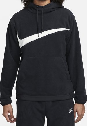 Osoba ubrana w czarną bluzę z kapturem Nike z dużym białym logo Swoosh na klatce piersiowej oraz pasujące czarne spodnie dresowe Nike z logo na lewej udzie.