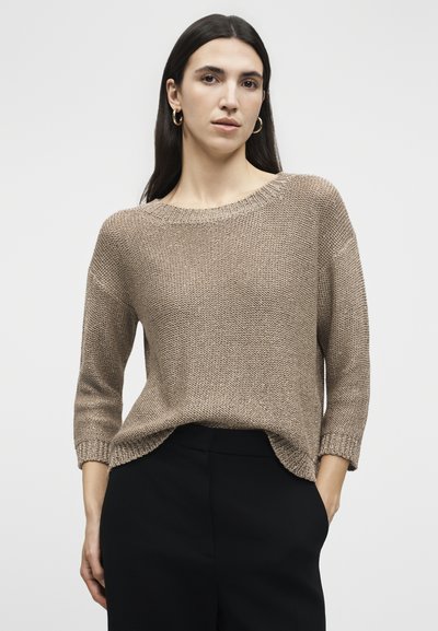 Femme aux longs cheveux foncés portant des boucles d'oreilles créoles dorées, un pull en maille marron ample à manches trois quarts et un pantalon noir les mains dans les poches.