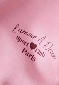 Tejido de algodón rosa con texto impreso: "L'amour À Deux Sport ♥ Club Paris" en fuentes cursivas y negritas marrones oscuras. Textura suave.