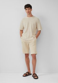 Beige gebreid T-shirt met korte mouwen en bijpassende korte broek, met een gestructureerd patroon. Gerd gedragen met bruine sandalen, handen in de zakken.