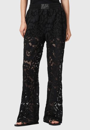 Schwarze transparente Spitzhosen mit Blumenmustern, getragen mit schwarzen Sandalen und einem schwarzen Oberteil, zeigen eine lockere Passform und einen elastischen Bund.
