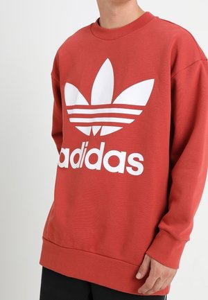 Osoba nosząca czerwoną bluzę Adidas z dużym białym logo trefoil i nazwą marki z przodu, stojąca na jednolitym białym tle.
