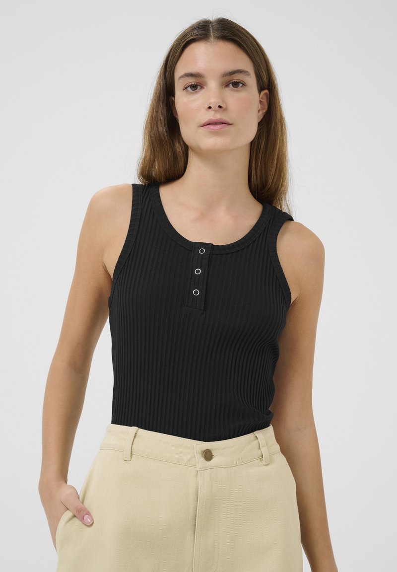 Top in cotone nero a coste con scollatura rotonda e patta con tre bottoni. Il tessuto è morbido e ha una forma aderente. Abbinato a pantaloni beige a vita alta.