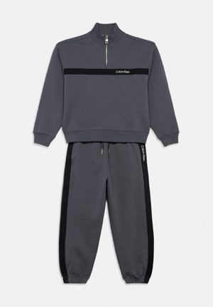 Ciemnoszary dres dwuczęściowy z półgolfem na suwak i elastycznymi mankietami, z czarnymi paskami i logo "Calvin Klein" na piersi oraz spodniach.