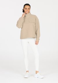 Beiger Fleece-Pullover mit hohem Kragen und Brusttasche; lockere Passform. Kombiniert mit enganliegenden weißen Leggings und weißen Turnschuhen.