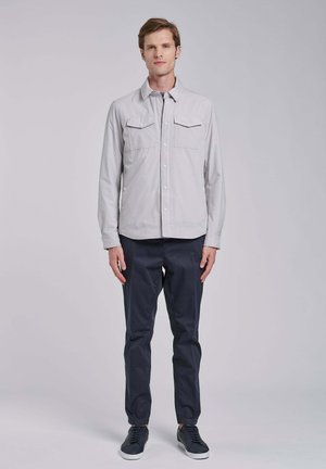 Man staand, dragen een lichtgrijze jas met knopen, donkerblauwe broek en zwarte sneakers tegen een effen grijze achtergrond.