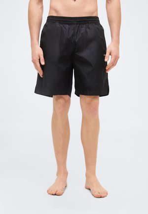 Mandlig model iført sorte midt-lange shorts med elastisk talje, stående barfodet mod en ensartet hvid baggrund.