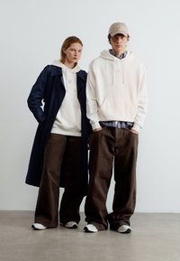 Νεαρός άνδρας και γυναίκα στέκονται δίπλα-δίπλα, και οι δύο φορούν λευκές κουκούλες, καφέ παντελόνια με φαρδιά μπατζάκια και λευκά αθλητικά παπούτσια· η γυναίκα φοράει επίσης ένα μακρύ σκοτεινό παλτό.