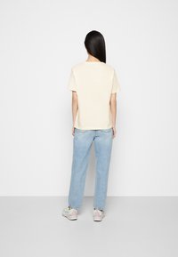 Mujer con cabello largo y negro, vistiendo una camiseta beige sencilla, jeans azul claro y zapatillas blancas, de pie frente a una pared blanca.