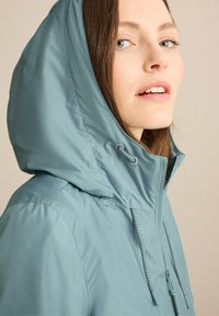 Veste à capuche bleu clair avec une texture lisse, dotée de cordons de serrage réglables, d'une fermeture éclair et d'un design ajusté au niveau du col.