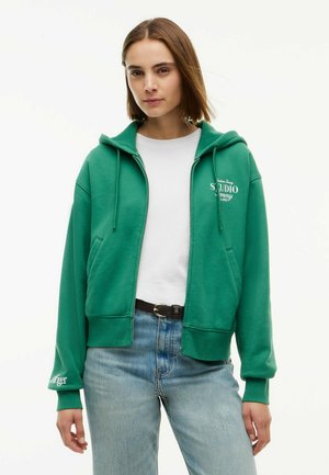 Ung kvinde iført en grøn lynlåshættetrøje over en hvid t-shirt og lyseblå jeans, stående mod en ensfarvet hvid baggrund.