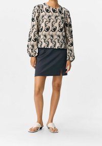 Femme portant un blouse beige à manches longues avec des broderies florales noires, une jupe noire avec une fente à l'avant, et des sandales ouvertes blanches.