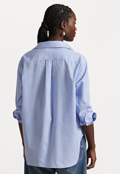 Femme aux cheveux tressés portant une chemise bleu clair à manches longues avec les manches retroussées, debout dos à un fond blanc.