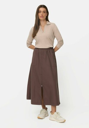 Kvinde står iført beige ribbet langærmet top, mørkebrun midi nederdel med lynlås foran og hvide sneakers på hvid baggrund.