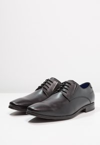 Zapatos de vestir de cuero negro con punta afilada, superficie lisa, cordones decorativos y un tacón bajo. Con un sutil relieve en la parte superior.