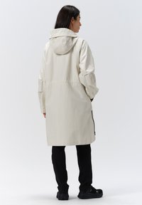 Giacca impermeabile leggera color beige con cappuccio, polsini elasticizzati e vita con cordino. La parte posteriore presenta un taglio lungo e tasche laterali con zip.