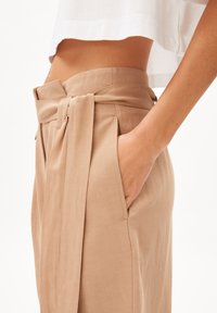 Beige pants featuring a tied waistband, side pockets, and a smooth, textured fabric. The design includes a relaxed fit and a high waist.