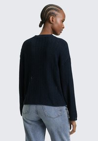 Vrouw met cornrows die een marineblauwe gebreide trui en lichtblauwe spijkerbroek draagt, staand en naar de zijkant kijkend tegen een effen achtergrond.