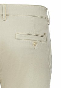 Beige bomuldsbukser med en baglomme, der har en knaplukning. Stoffet har en tekstureret overflade med diskrete syninger.