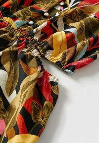 Tissu coloré avec des motifs abstraits de feuilles rouges, jaunes, brunes et noires, avec des poignets élastiqués et trois boutons blancs sur une manche.