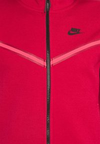 Röd zip-up jacka med svart dragkedja, prydd med en svart Nike-logotyp och kontrasterande rosa linjer över bröstet. Mjuk tyg, sportig design.