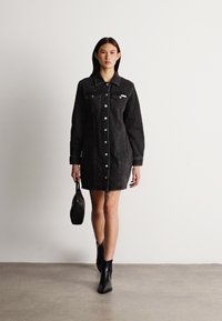 Vestido camisola de ganga preta com mangas longas, frente abotoada e dois bolsos no peito. Estilizado com botas ankle pretas e uma pequena mala.