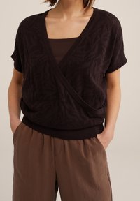 Tmavě hnědý pletený top s výstřihem do V a krátkými rukávy, zdobený texturovaným vzorem. Kombinován s volnými, světle hnědými kalhotami.