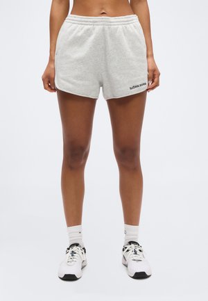 Kvinde iført lysegrå Björn Borg-shorts, hvide sokker og hvide sneakers, stående mod en ensfarvet lys baggrund.