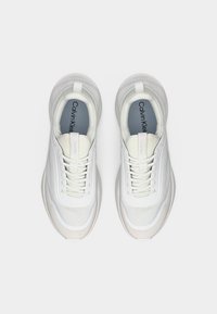 Ζευγάρι λευκών αθλητικών παπουτσιών Calvin Klein με λεπτομέρειες από δίχτυ και δέρμα, ορατά κορδόνια και επώνυμα εσωτερικά πέλματα, σε λευκό φόντο, από ψηλά.