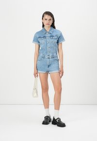 Salopette en denim bleu clair avec des manches courtes, un devant à boutons et un short retroussé. Assortie avec des chaussures noires et un petit sac à main blanc.