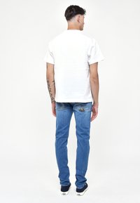 T-shirt bianco a girocollo, jeans in denim blu e sneaker blu navy. Design semplice con maniche corte e vestibilità dritta. Tatuaggio visibile sul braccio.