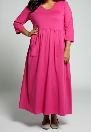 Femme portant une longue robe rose vif avec des manches trois-quarts et deux poches avant, debout avec une main sur la hanche et des chaussures beiges.