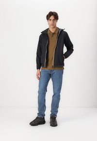 Black hooded jacket with elastic cuffs, front zipper, and yellow stripe; paired with a brown polo, blue jeans, and dark sneakers.