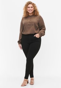 Camisa de manga larga marrón con detalles de botones y pliegues, combinada con pantalones negros ajustados. La modelo lleva sandalias de tiras marrón claro. Fondo simple.