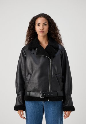 Veste en simili cuir noir avec un col en fausse fourrure, une taille ceinturée et des poignets texturés. Comprend une fermeture éclair frontale et des poches latérales.