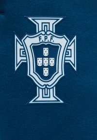 Έμβλημα της Ποδοσφαιρικής Ομοσπονδίας Πορτογαλίας με λευκό σταυρό και ασπίδα που περιλαμβάνει πέντε μικρότερες ασπίδες με κουκκίδες σε σκούρο μπλε φόντο από ύφασμα.