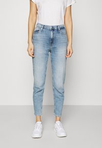 Světle modré džíny s vysokým pasem a zúženými nohavicemi, vyrobené z denimové látky. Obsahují přední kapsy a minimální švy. Nosí se s bílými teniskami.