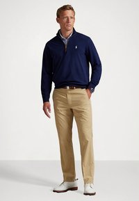 Marineblå genser med glidelås og logo, kombinert med kaki chinos og hvite sko. Antrekket inkluderer en lagvis blå skjorte med krage under genseren.