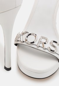 Sandale blanche à talon haut avec semelle plateforme et lettres en métal argenté formant "KORS" sur la bride avant.